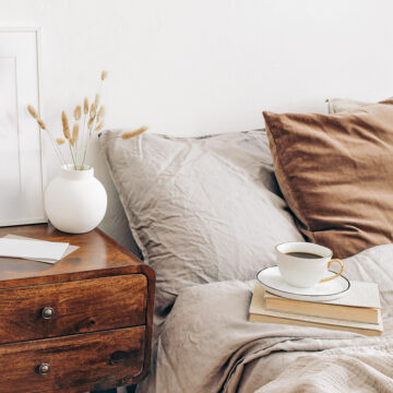 Portrait white frame mockup on retro wooden bedside table. Modern white ceramic vase, dry grass. Cup of coffee and books in bed. Beige linen pillows in bedroom. Scandinavian interior.